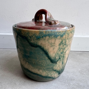 May include: A ceramic jar with a lid, featuring a unique design. The jar's exterior showcases a blend of teal, beige, and brown hues, with a textured, crackled effect. The lid is a deep red with a curved handle, adding a decorative touch.