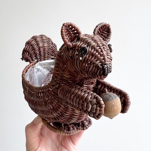May include: A brown wicker squirrel-shaped basket with a clear plastic liner. The squirrel holds a wooden acorn. The basket is designed with woven details, including the squirrel's ears, tail, and paws. A decorative item for home decor.