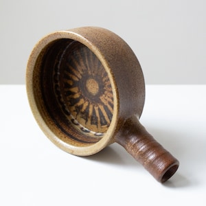 Könnte beinhalten: Eine braune Keramikschüssel mit einem braunen und weißen geometrischen Muster im Inneren. Die Schüssel hat einen Griff an der Seite.