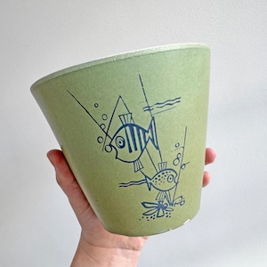 May include: A light green, vintage-style planter with a tapered shape. The planter features a blue illustration of two fish, bubbles, and aquatic plants. The design is reminiscent of mid-century modern art. The planter is held by a hand.