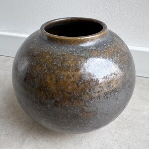 Pode incluir: Um vaso de cerâmica grande e redondo com um acabamento escuro e manchado. O vaso tem uma abertura larga e uma superfície lisa e brilhante. As cores variam do castanho escuro ao bronze e ao cinzento, criando uma aparência texturizada. Uma peça decorativa para decoração de casa.