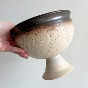 May include: A handmade ceramic bowl with a pedestal base. The bowl has a textured, off-white exterior with a dark brown rim. The bowl is held in a hand.
