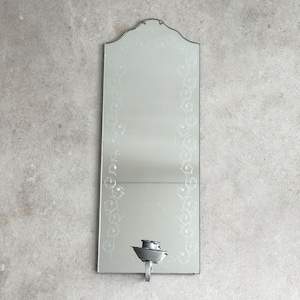 Może przedstawiać: Prostokątne lustro ścienne z dekoracyjną górną krawędzią i wytrawionymi wzorami kwiatowymi po bokach. Mały, srebrny świecznik jest przymocowany u dołu. Lustro jest zamontowane na jasnoszarej ścianie.