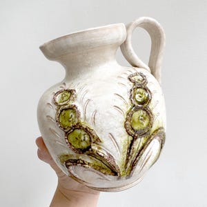 Op de afbeelding: Een crèmekleurige keramische kan met een wijde opening en een gebogen handvat. Het lichaam is voorzien van reliëf, gestructureerde bloemmotieven in olijfgroene en bruine tinten, met uitgesneden bladachtige patronen rond de centrale elementen.
