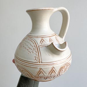 May include: Cream-coloured ceramic jug with a handle and spout. The jug has embossed geometric patterns in a darker cream shade, including triangles, swirls, and textured areas. The design gives it a rustic, handcrafted appearance.