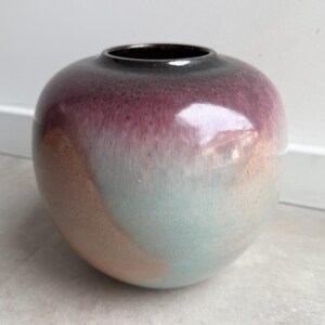 May include: A round ceramic vase with a smooth, glossy finish. The upper portion features a deep plum purple color that transitions into a soft, muted teal and a warm, earthy peach tone toward the base. The vase has a small, circular opening.