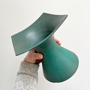 Puede incluir: Un jarrón de cerámica de color verde azulado con una parte superior única y acampanada y una base redondeada. El jarrón está sostenido por una persona que lleva un suéter gris. El jarrón tiene un acabado mate y una superficie texturizada.