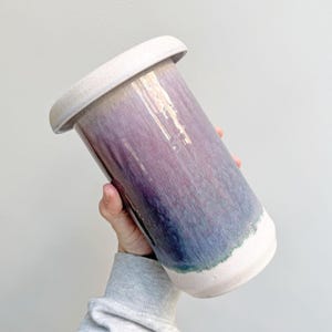 Può includere: Un vaso cilindrico in ceramica con un bordo largo, caratterizzato da una sfumatura di colori. La parte superiore e inferiore sono beige chiaro, mentre il centro passa dal viola al blu. Il vaso è tenuto ad angolo.