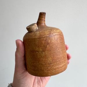Könnte beinhalten: Eine kleine, braune Keramikflasche mit einem Korkstopfen. Die Flasche hat eine strukturierte Oberfläche und eine abgerundete Form.