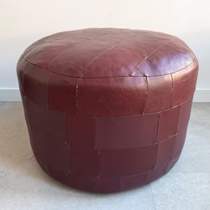 Puede incluir: Un otomán de cuero burdeos con una parte superior redondeada y una base cilíndrica. El otomán está construido con piezas rectangulares de cuero cosidas, creando una superficie texturizada. Es un mueble decorativo.
