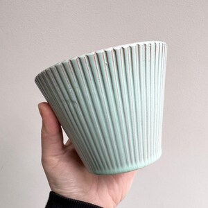 May include: A pale blue ceramic plant pot with vertical ribbed detailing. The pot has a slightly flared shape and a textured surface. The rim of the pot has a contrasting brown colour. The pot is held in a hand.