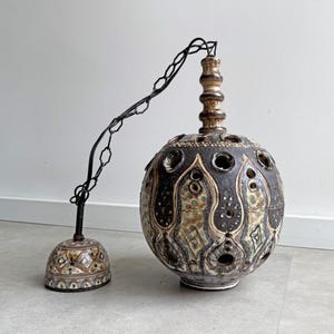 Peut inclure: Un luminaire suspendu en céramique brun et blanc avec un motif géométrique et des trous. La lumière est suspendue à une chaîne noire.