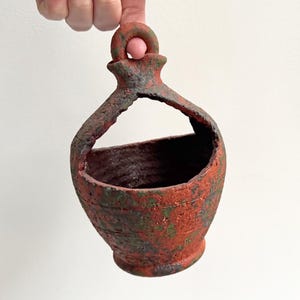 Könnte beinhalten: Ein rustikaler Keramik-Hängepflanztopf mit rot-grüner, gesprenkelter Oberfläche. Der Pflanztopf hat einen runden Körper, eine breite Öffnung und einen Henkel zum Aufhängen. Die Textur deutet auf ein gealtertes, verwittertes Aussehen hin.