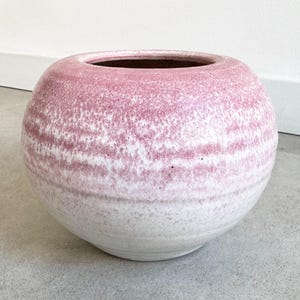 May include: A round, ceramic vase with a speckled pink and white exterior. The vase has a wide opening and a smooth, rounded shape. The color gradient transitions from white at the base to pink at the top. The vase is suitable for home decor.