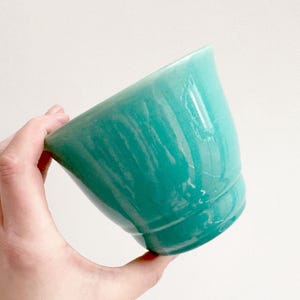 Può includere: Una ciotola in ceramica turchese con finitura lucida. La ciotola ha un bordo leggermente svasato e una base arrotondata. Il colore è un verde acqua vibrante e la superficie riflette la luce, evidenziando la consistenza della smaltatura.