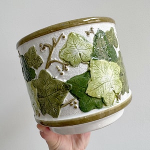 Op de afbeelding: Witte keramische plantenpot met een reliëfontwerp van groene klimopbladeren en -ranken. De rand en de basis zijn afgewerkt met een donkergroene band. De pot is cilindrisch en heeft een licht uitlopende basis.