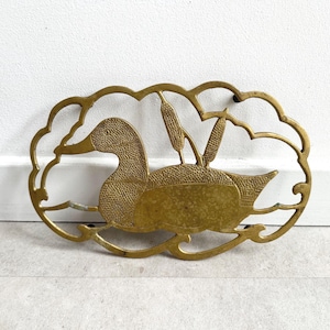 May include: A brass duck wall hanging. The duck is depicted swimming with cattails in the background. The brass is a warm gold color. The duck is inside an oval frame with a wavy edge.