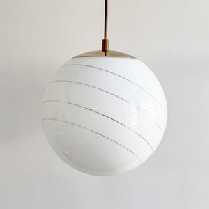 Puede incluir: Una l&aacute;mpara colgante de globo blanco con rayas doradas. La luz cuelga de un techo blanco.