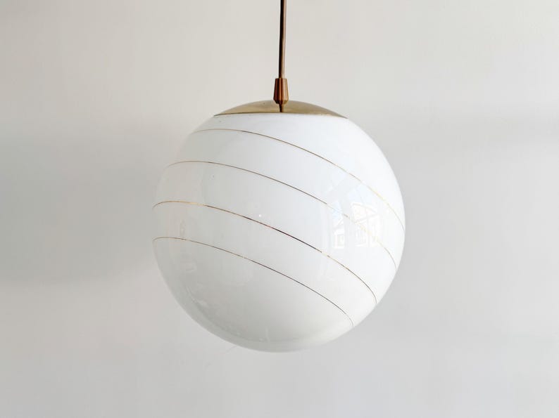 Puede incluir: Una l&aacute;mpara colgante de globo blanco con rayas doradas. La luz cuelga de un techo blanco.