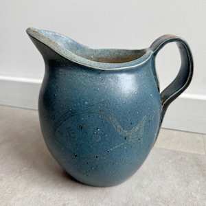Op de afbeelding: Een blauwe keramische kan met een gebogen handvat en een tuit. De kan heeft een gespikkelde textuur en een ontwerp dat in de zijkant is geëtst. De kan heeft een gedempte blauwe kleur en is geschikt voor het serveren van dranken.