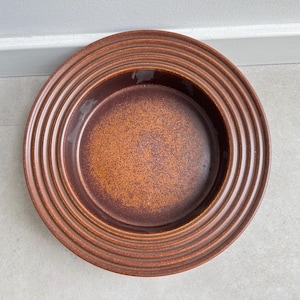 May include: A brown ceramic plate with a textured, ridged rim. The center of the plate has a speckled, orange-brown appearance. The plate is circular and appears to be a serving dish.
