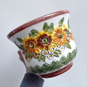 Könnte beinhalten: Weißer Keramik-Blumentopf mit floralem Design. Der Topf hat erhabene, farbenfrohe Blumen in Gelb-, Orange- und Rottönen mit grünen Blättern. Der Rand und der Boden sind dunkelrot.
