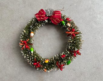 May include: A festive Christmas wreath with a green base, adorned with red berries, colorful lights, and a red bow with gold stripes. The wreath is circular and appears to be made of artificial evergreen branches.