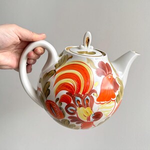 Könnte beinhalten: Weiße Keramik-Teekanne mit großem Henkel und Deckel. Die Teekanne ist mit einem farbenfrohen Hahn-Design in Orange-, Rot-, Gelb- und Brauntönen verziert. Das Design ist handbemalt mit floralen Akzenten.
