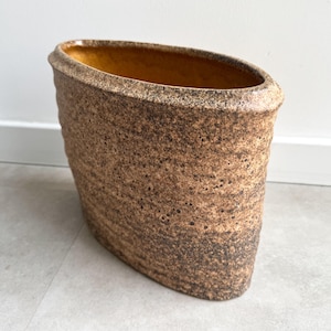 May include: A brown, oval-shaped ceramic vase with a textured surface. The vase has a wide opening and a slightly flared rim. The interior is a warm, golden color. The exterior has a speckled, earthy appearance, suggesting a handcrafted aesthetic.