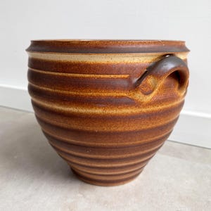 Puede incluir: Una maceta de cerámica marrón hecha a mano, con un cuerpo ancho y acanalado y dos asas curvas. La maceta tiene un borde de color beige claro y una base marrón oscuro, con una transición gradual de color. Adecuada para plantas o almacenamiento.