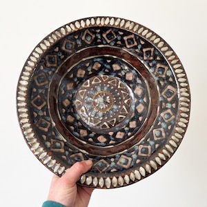 May include: A large, round ceramic bowl with a brown, black and white geometric pattern. The bowl has a raised rim with a white, scalloped edge.