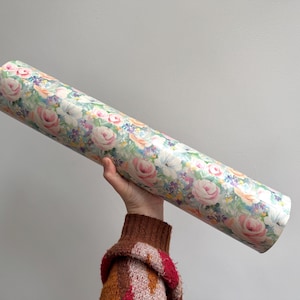 Könnte beinhalten: Eine Rolle floral gemustertes Papier mit rosa Rosen, weißen Blumen und anderen bunten Blüten. Das Papier wird von einer Person gehalten, die einen braunen und roten Pullover trägt. Der Hintergrund ist einfarbig hellgrau.