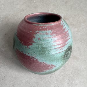 May include: A round ceramic vase with a wide opening. The vase is painted with a blend of red and turquoise colors, creating a textured, artistic appearance. The interior is dark. This decorative piece is suitable for home decor.