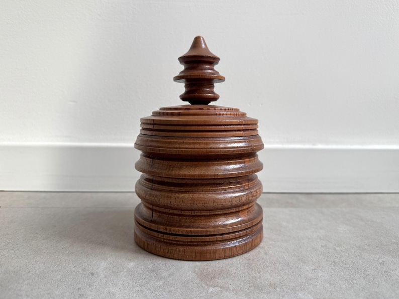 Pu&ograve; includere: Una scatola di legno con un design a strati e a gradini e una parte superiore appuntita. La scatola &egrave; realizzata in legno marrone scuro e ha una finitura liscia e lucida.