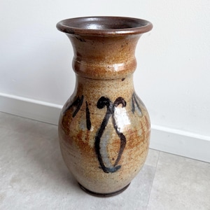 May include: A handcrafted ceramic vase with a wide base and a flared rim. The vase features a speckled tan and brown glaze with dark brown and blue abstract designs. The vase is approximately 12 inches tall.