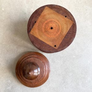 Pu&ograve; includere: Una scatola di legno con un coperchio. Il coperchio ha un design quadrato al centro con un foro circolare. La scatola &egrave; in legno scuro con una macchia marrone chiaro.