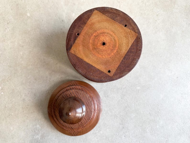 Pu&ograve; includere: Una scatola di legno con un coperchio. Il coperchio ha un design quadrato al centro con un foro circolare. La scatola &egrave; in legno scuro con una macchia marrone chiaro.