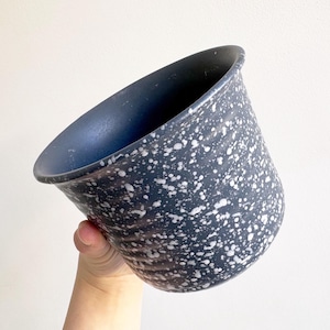 May include: A speckled, dark gray ceramic planter with a flared rim. The planter has a speckled pattern of white dots across its surface. The interior is a solid dark gray. The planter is held against a white background.