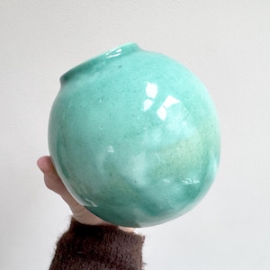 Puede incluir: Un jarrón redondo de cerámica turquesa con acabado brillante. El jarrón tiene una pequeña abertura en la parte superior y lo sostiene una persona. El jarrón es un objeto decorativo.