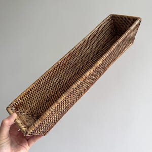 Peut inclure: Un plateau rectangulaire long et étroit fabriqué en rotin tressé marron. Le plateau présente un motif de tissage serré et uniforme sur toute sa structure, avec des côtés légèrement évasés et une base plate, offrant un aspect naturel texturé.