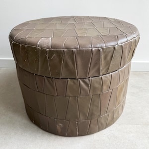 May include: A round, taupe-colored ottoman featuring a segmented, geometric pattern of stitched panels covering the entire exterior surface, including the flat top and the cylindrical base, with visible signs of wear on the leather-like material.