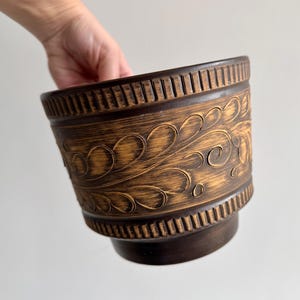 Op de afbeelding: Een bruine keramische bloempot met een decoratief bloemmotief. De pot heeft een brede, cilindrische vorm met een licht uitlopende basis. De buitenkant heeft een gestructureerd, gesneden ontwerp.
