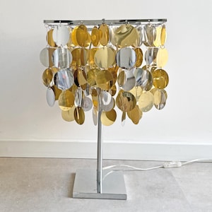 May include: A table lamp featuring a square chrome base and a central chrome stem supporting a square frame. The shade consists of multiple hanging circular discs in alternating gold and silver metallic finishes, suspended by small metal rings.
