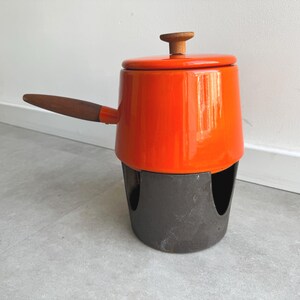 Könnte beinhalten: Ein leuchtend orangefarbener Emailletopf mit einem Holzgriff und einem passenden Deckel mit kleinem Holzknauf. Der Topf steht sicher in einem dunkelgrauen, zylindrischen Metallständer mit großen bogenförmigen Aussparungen am Boden.