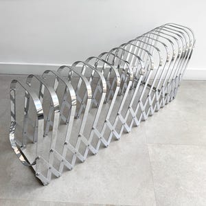 May include: A chrome-plated metal magazine rack featuring an expandable accordion-style base with multiple vertical U-shaped dividers. The structure is composed of polished silver-toned metal segments connected by a series of pivoting joints.