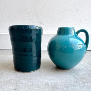 Könnte beinhalten: Zwei Keramikgefäße. Links ein dunkelblaugrüner, zylindrischer Becher mit unebenem Rand. Rechts eine türkisfarbene, runde Kanne mit Henkel. Beide stehen auf einer grauen Oberfläche vor einer weißen Wand.