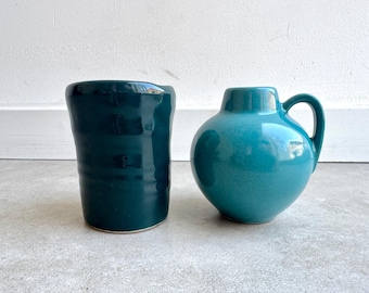 May include: Two ceramic vessels. On the left, a dark teal, cylindrical cup with a slightly uneven rim. On the right, a turquoise, round jug with a handle. Both are on a gray surface, against a white wall.
