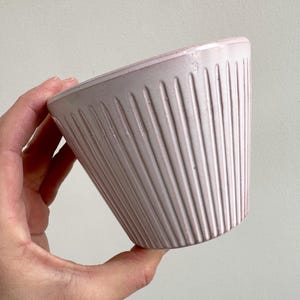 Op de afbeelding: Een lichtroze keramische koffiefilterhouder met verticale geribbelde details. De houder wordt door een hand vastgehouden en is naar de kijker toe gekanteld. De houder is lichtroze van kleur en heeft een gladde, licht glanzende afwerking.