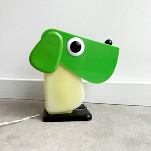 Könnte beinhalten: Eine grüne und gelbe Tischlampe aus Plastik in Form eines Hundes mit schwarzem Sockel. Die Lampe hat ein großes, rundes Auge und eine kleine, schwarze Nase.