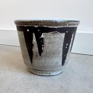 Op de afbeelding: Een keramische plantenpot met een witte glazuur en een zwart abstract ontwerp. De pot heeft een ronde vorm en een brede opening.